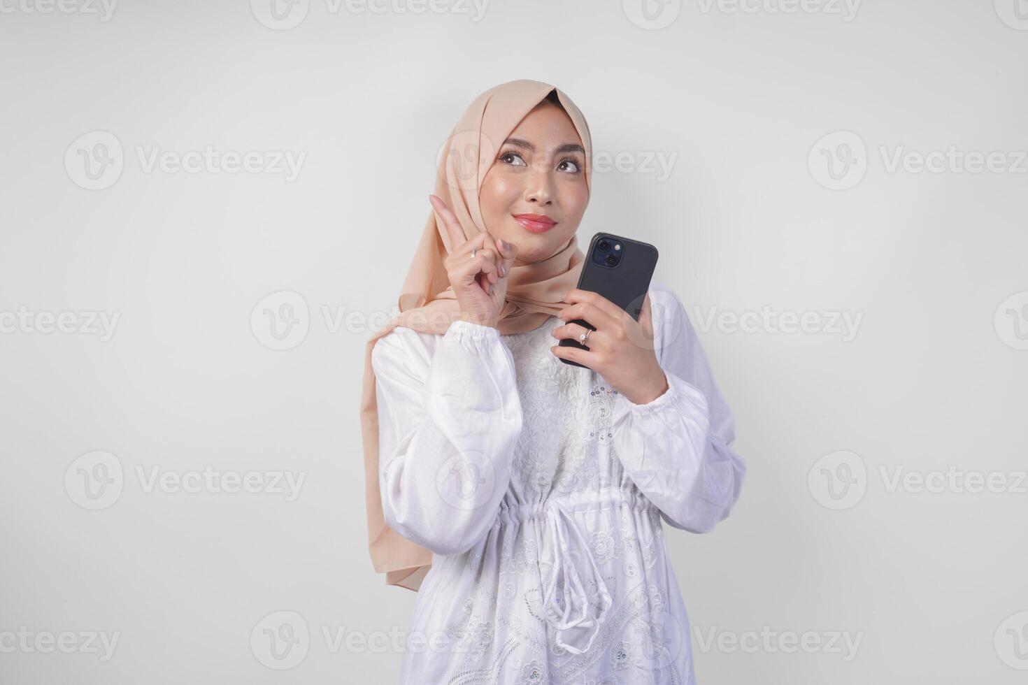 Thoughtful young Asian Muslim woman wearing white dress and hijab, using smartphone while holding her chin and thinking with serious expression over isolated white background photo