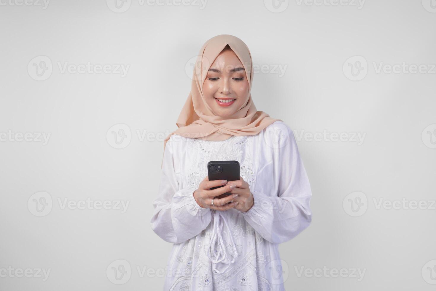 emocionado asiático musulmán mujer vistiendo blanco vestir y hijab participación su teléfono inteligente mientras sonriente alegremente, aislado por blanco antecedentes. ramadhan y eid Mubarak concepto foto
