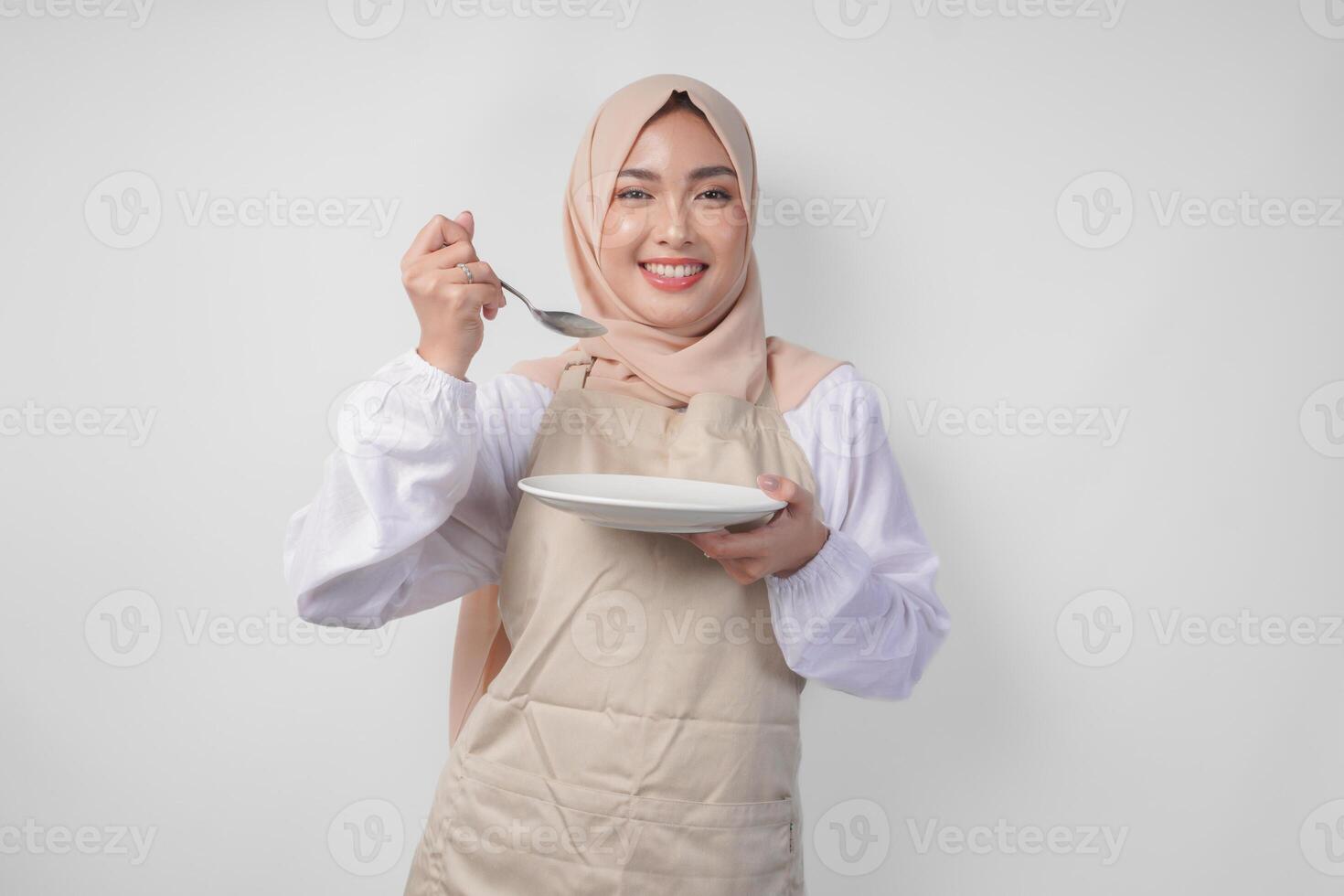 Hungry young Asian Muslim woman in hijab and cream apron holding spoon and eating from an empty plate with copy space over it. Ramadan concept photo