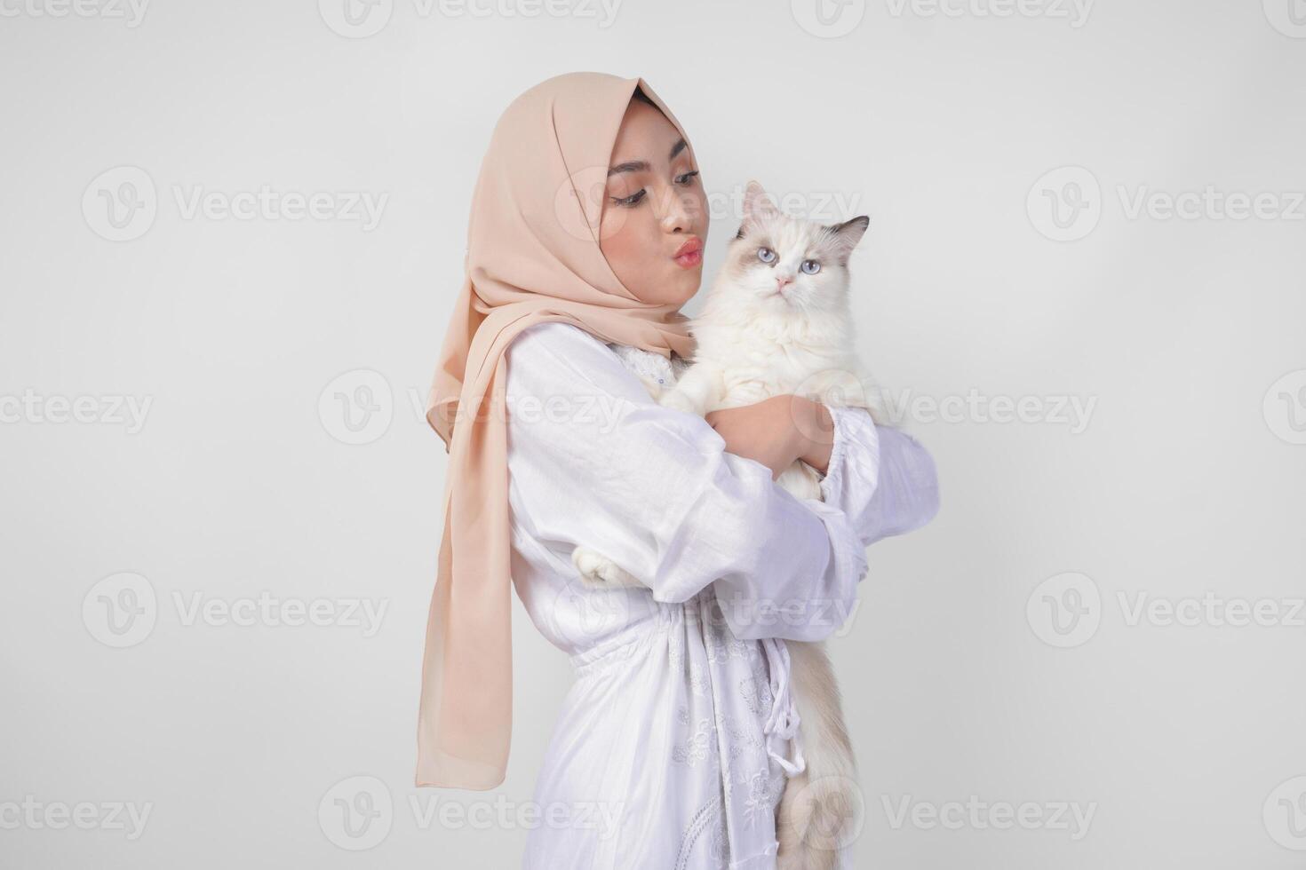 retrato de un hermosa joven musulmán mujer vistiendo blanco vestir y hiyab, sonriente y participación un blanco muñeca de trapo gato terminado aislado blanco antecedentes foto