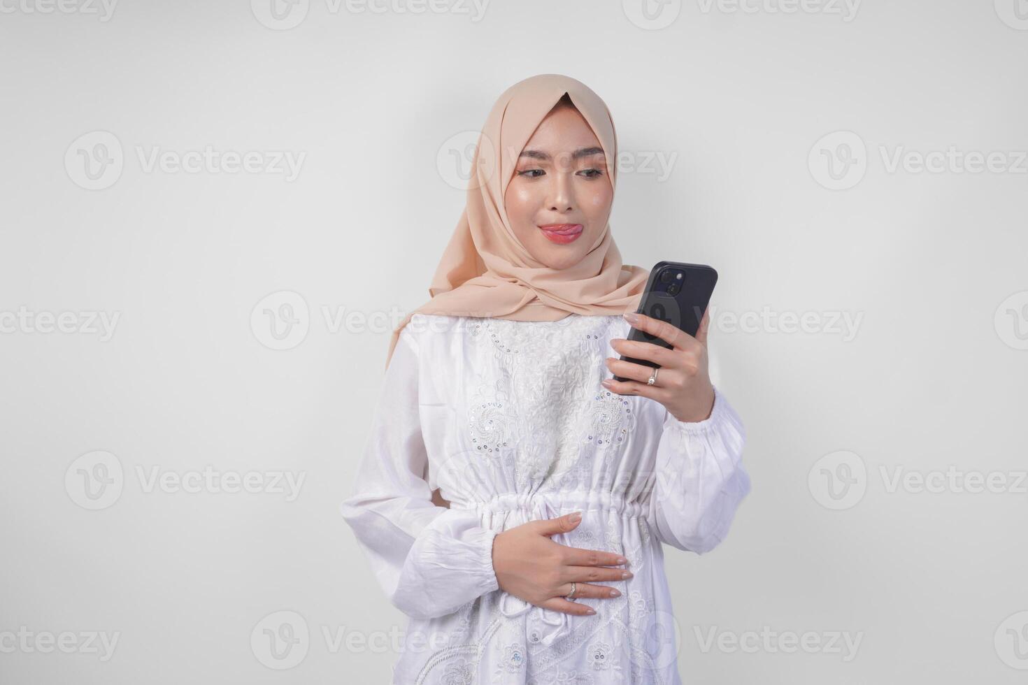 retrato de joven asiático musulmán mujer en blanco vestir y hijab sensación hambriento mientras mirando a su teléfono a orden sabroso comida después ayuno, aislado en blanco antecedentes foto