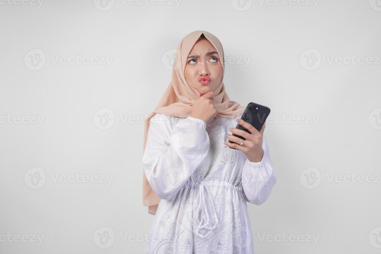 Thoughtful young Asian Muslim woman wearing white dress and hijab, using smartphone while holding her chin and thinking with serious expression over isolated white background photo