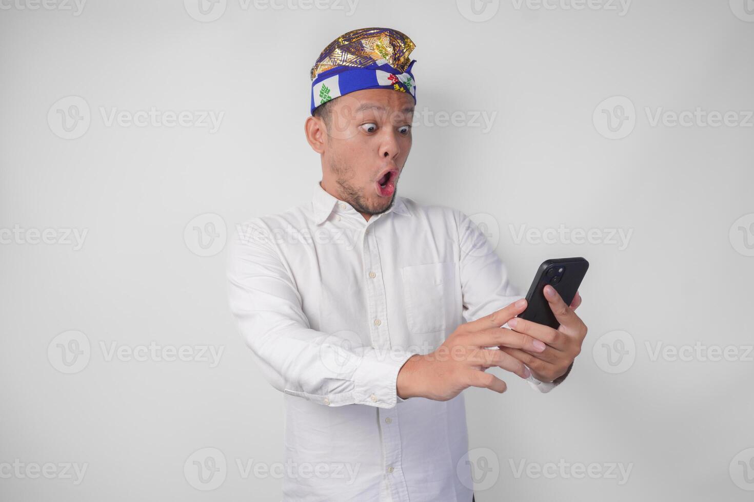 Young Balinese man wearing white shirt and traditional headdress showing shocked expression on face while holding smartphone, surprised after reading news or gossip, isolated by white background photo