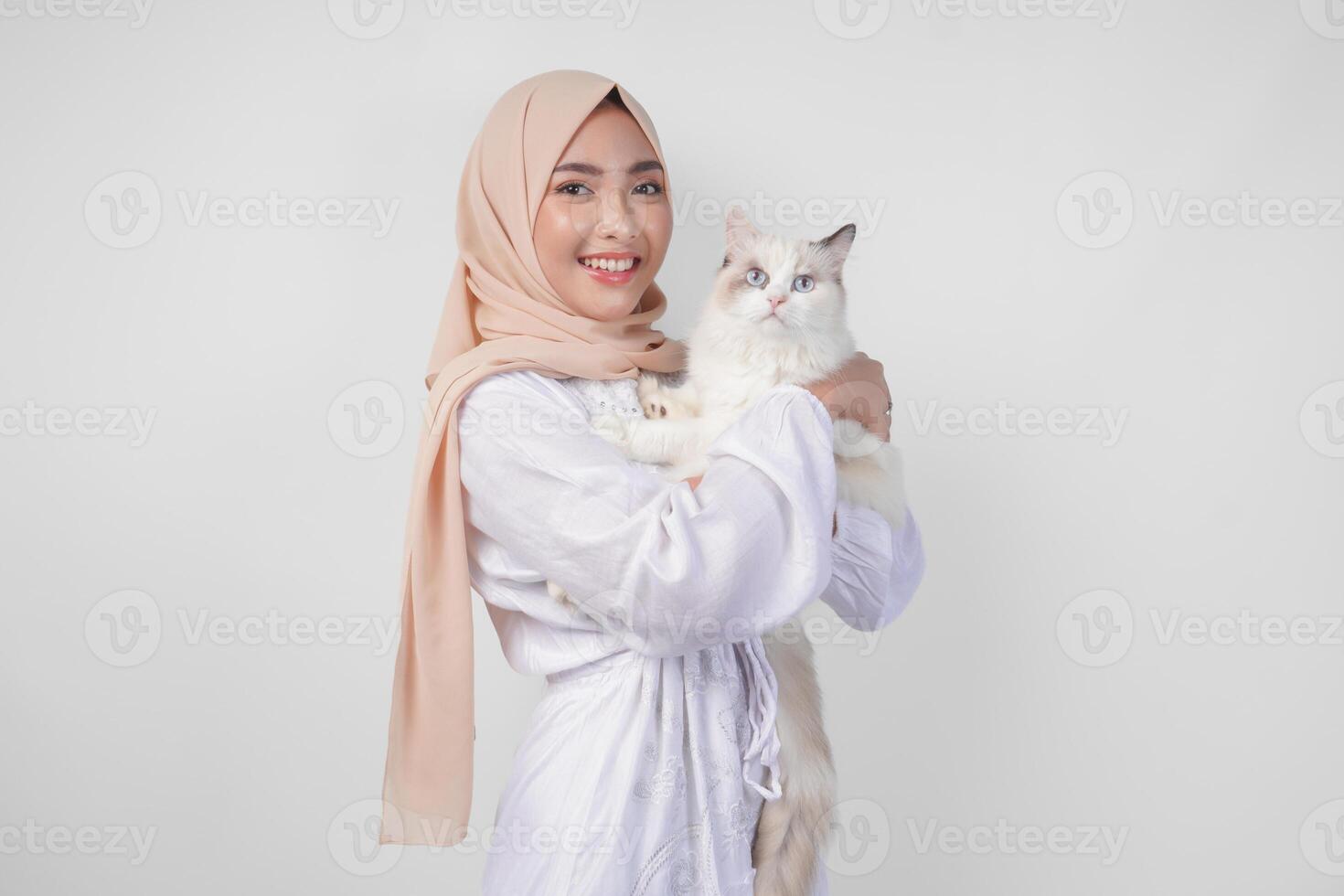 Portrait of a beautiful young Muslim woman wearing white dress and hijab, smiling and holding a white ragdoll cat over isolated white background photo