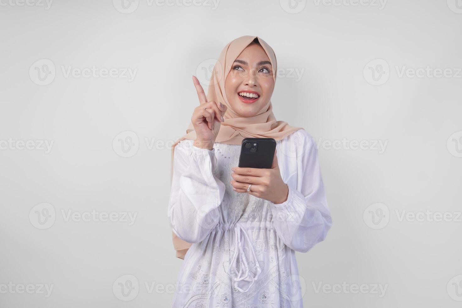 pensativo joven asiático musulmán mujer vistiendo blanco vestir y hijab participación teléfono mientras pensando y hallazgo idea con contento expresión, aislado en blanco antecedentes. Ramadán y eid Mubarak concepto foto