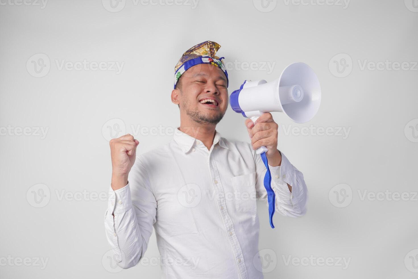 Energetic Balinese man in white shirt and traditional headdress shouting at megaphone while raising clenched fist, isolated by white background photo