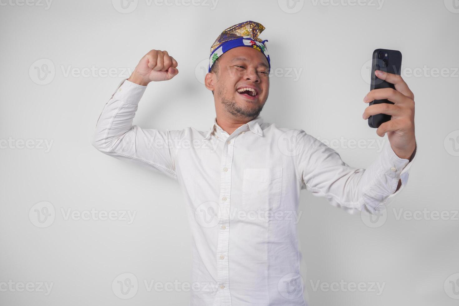 Young Balinese man in white shirt and traditional headdress doing call with family or friend feeling overjoyed and excited photo
