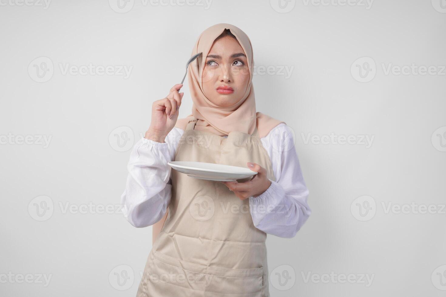 Confused young Asian Muslim woman in hijab and cream apron holding spoon and empty plate with copy space over it, thinking hard what food menu to eat for iftar. Ramadan concept photo