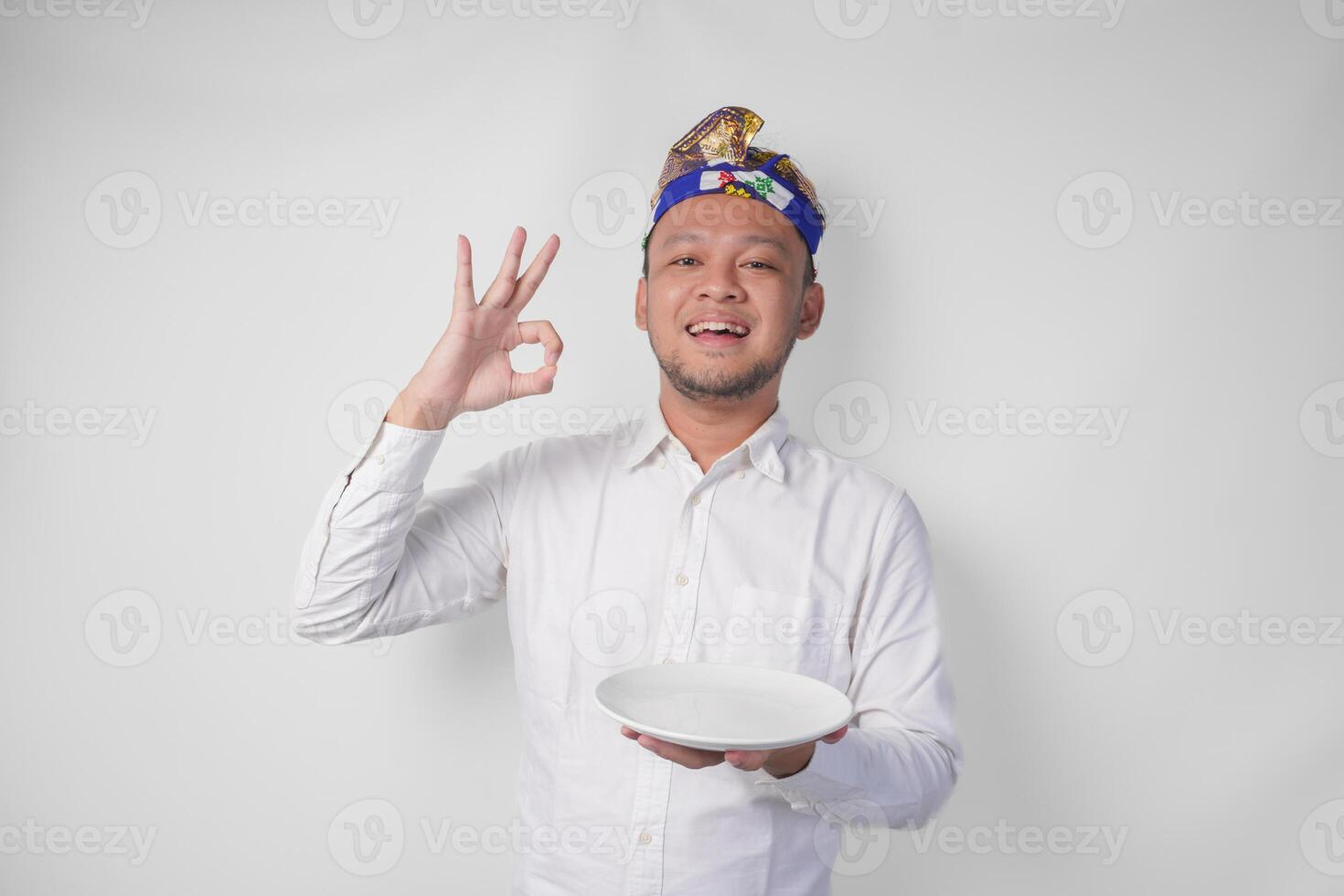 Happy young Balinese man in white shirt and traditional headdress holding an empty plate with copy space while making a delicious hand gesture to express how good the food is photo