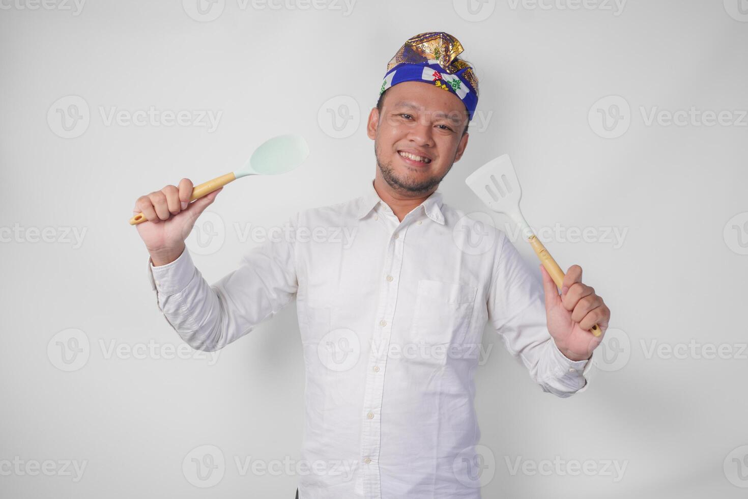 Smiling young Balinese man in white shirt and traditional headdress holding spatula and kitchen cooking utensils photo