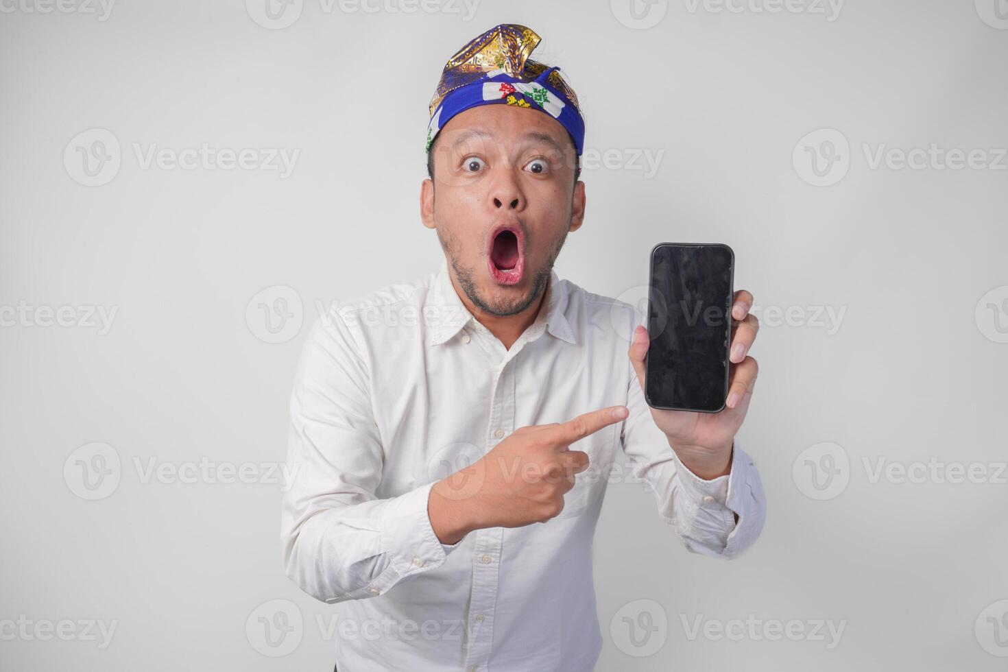 Surprised young Balinese man wearing white shirt and traditional headdress with shocked expression on face while pointing to his smartphone, presenting blank screen copy space photo