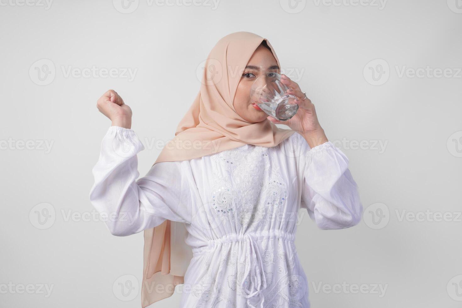 alegre asiático musulmán mujer vistiendo blanco vestir y hijab Bebiendo un vaso de agua con un contento exitoso expresión y gesto, sensación aliviado después ayuno, aislado por blanco antecedentes. foto