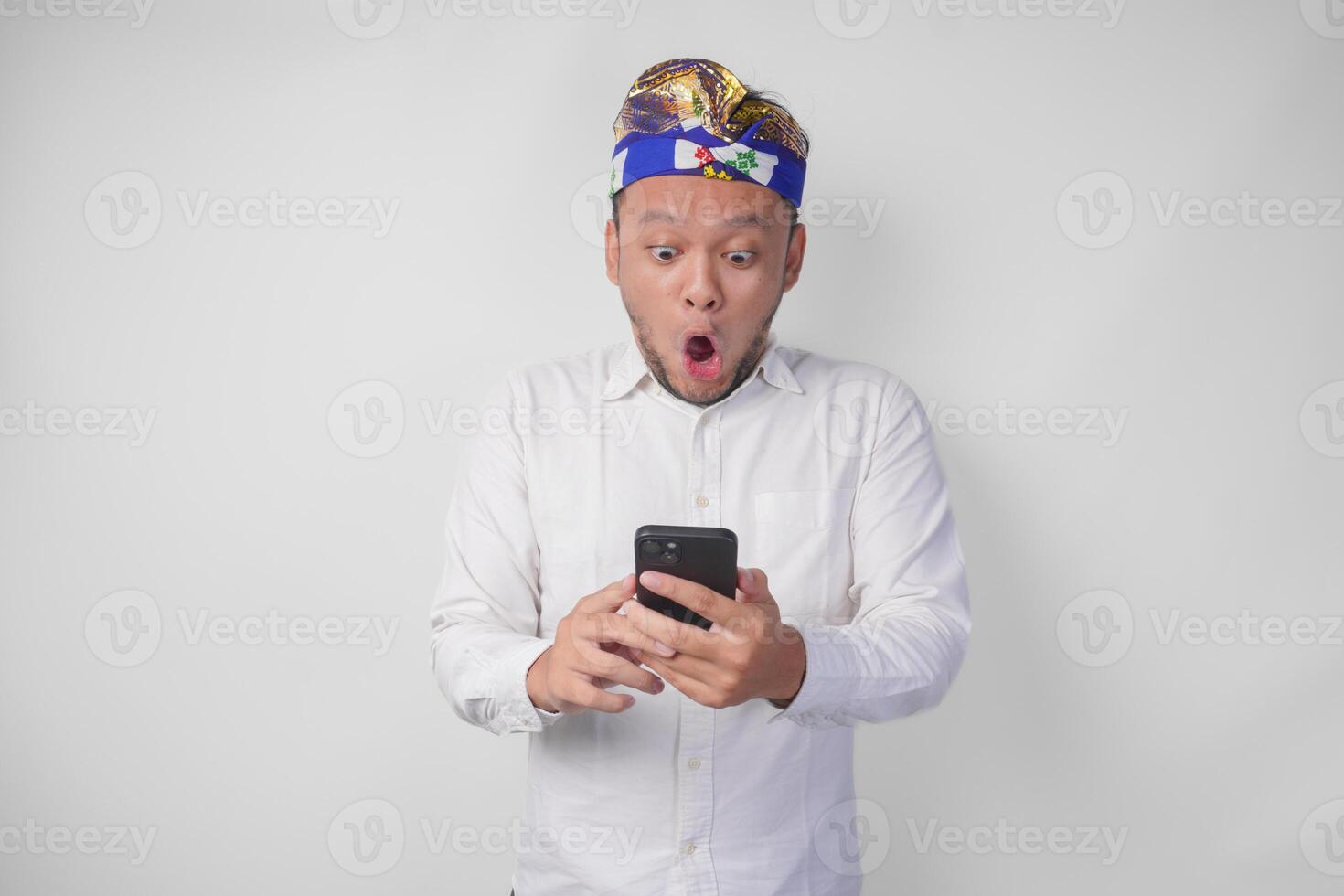 Young Balinese man wearing white shirt and traditional headdress showing shocked expression on face while holding smartphone, surprised after reading news or gossip, isolated by white background photo