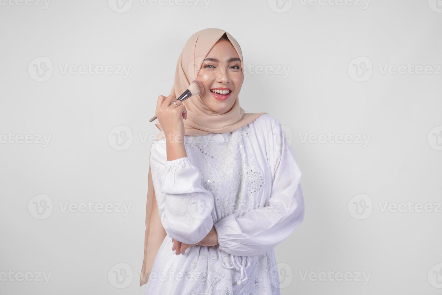 Beautiful young Asian Muslim woman wearing white dress and hijab applying make up using brush. Fashion and cosmetics beauty concept photo