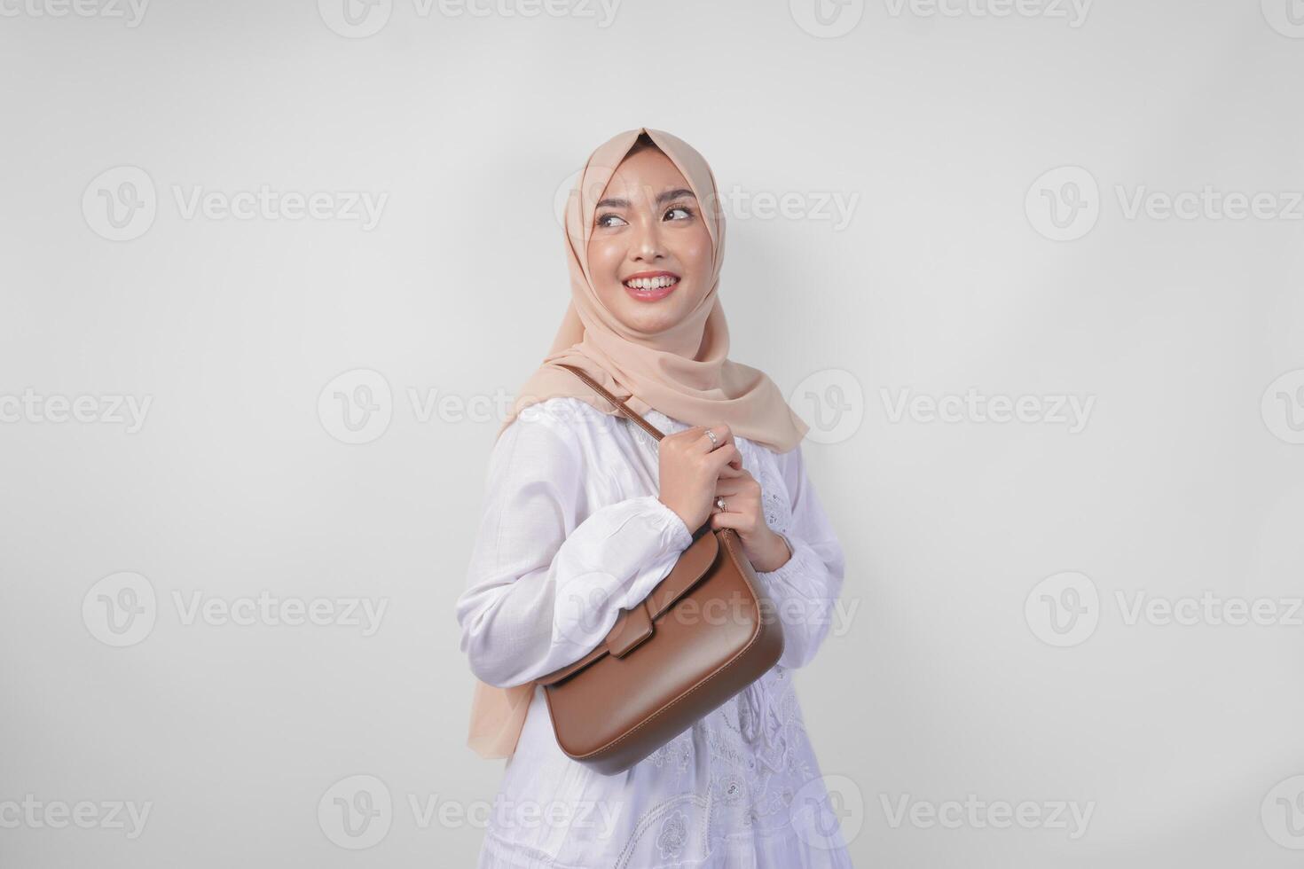 elegante joven asiático musulmán mujer vistiendo blanco vestir y hijab con marrón honda bolso sonriente felizmente terminado aislado blanco antecedentes. ramadhan Moda concepto foto