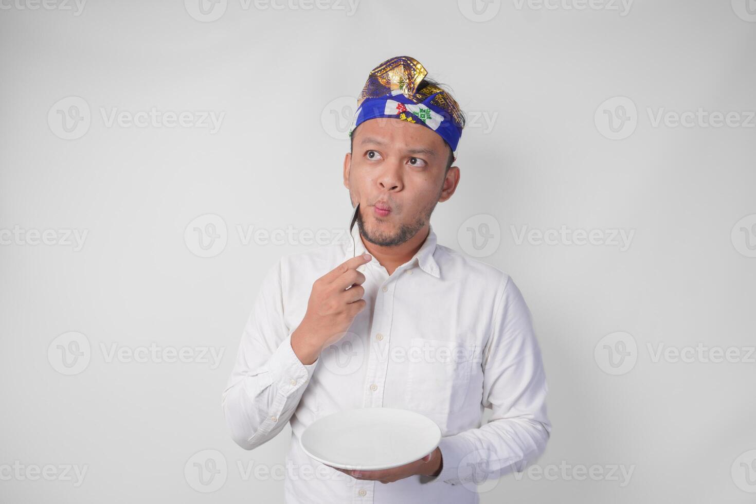 Thoughtful young Balinese man in white shirt and traditional headdress holding an empty plate with copy space and spoon while thinking what food to eat photo