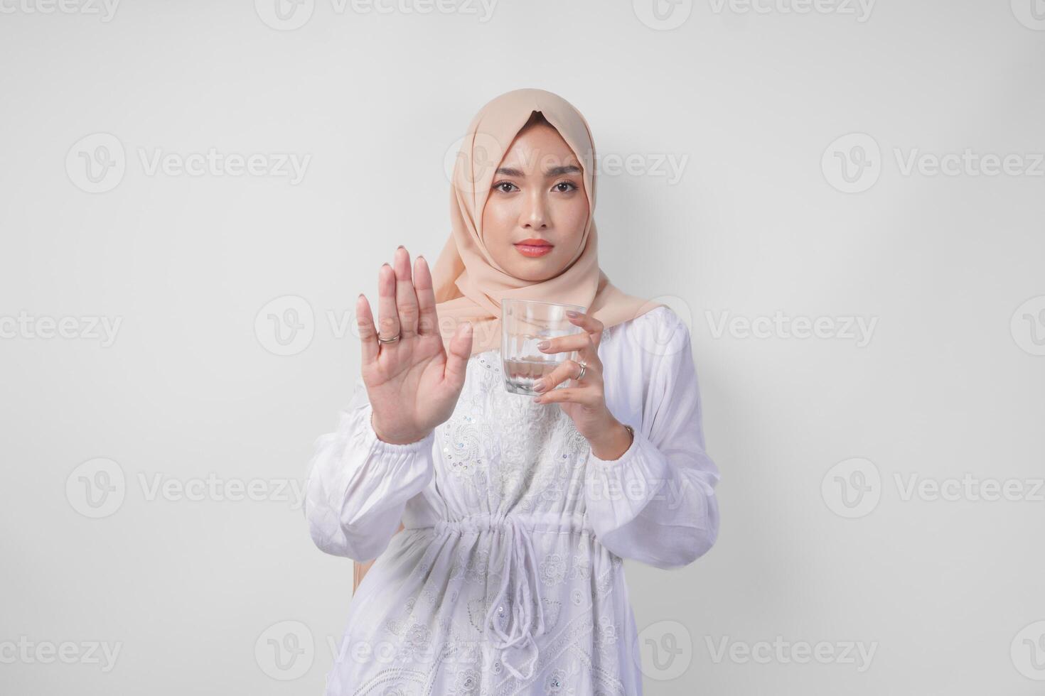 grave joven asiático musulmán mujer vistiendo blanco vestir y hijab demostración su palma a hacer un detener gesto mientras participación un taza de agua, en pie terminado aislado blanco antecedentes. Ramadán concepto foto