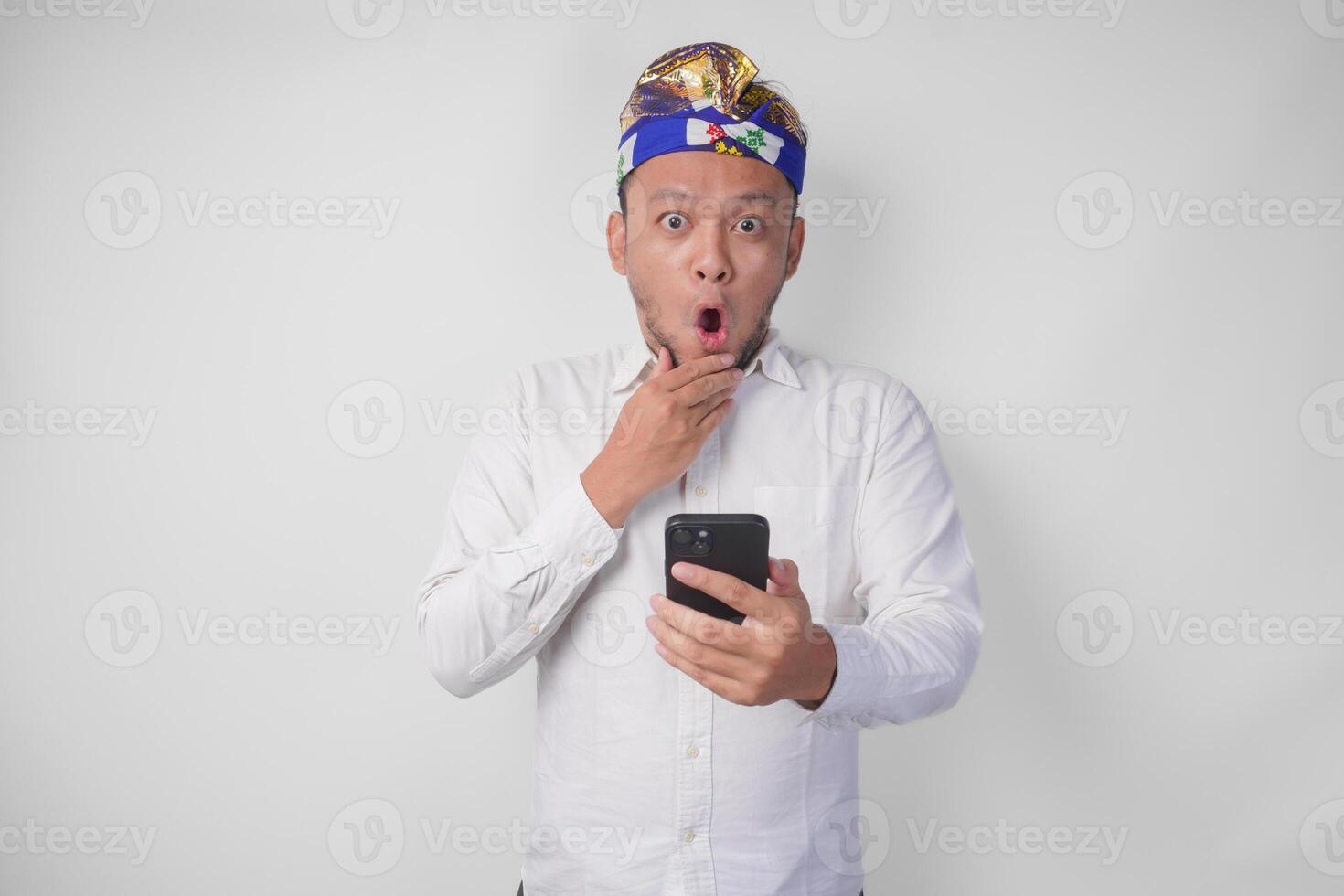 Young Balinese man wearing white shirt and traditional headdress showing shocked expression on face while holding smartphone, surprised after reading news or gossip, isolated by white background photo
