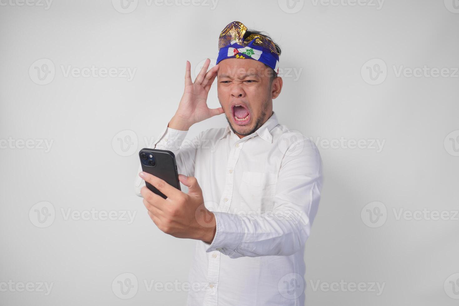 Angry Balinese man in white shirt and traditional headdress feeling mad and upset while using smartphone photo