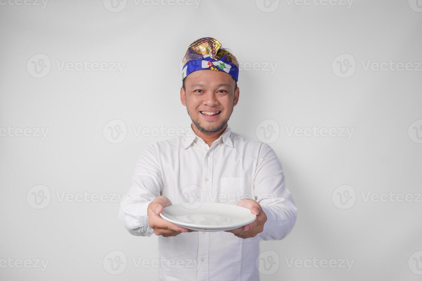 Smiling young Balinese man in white shirt and traditional headdress showing and presenting an empty plate with copy space photo