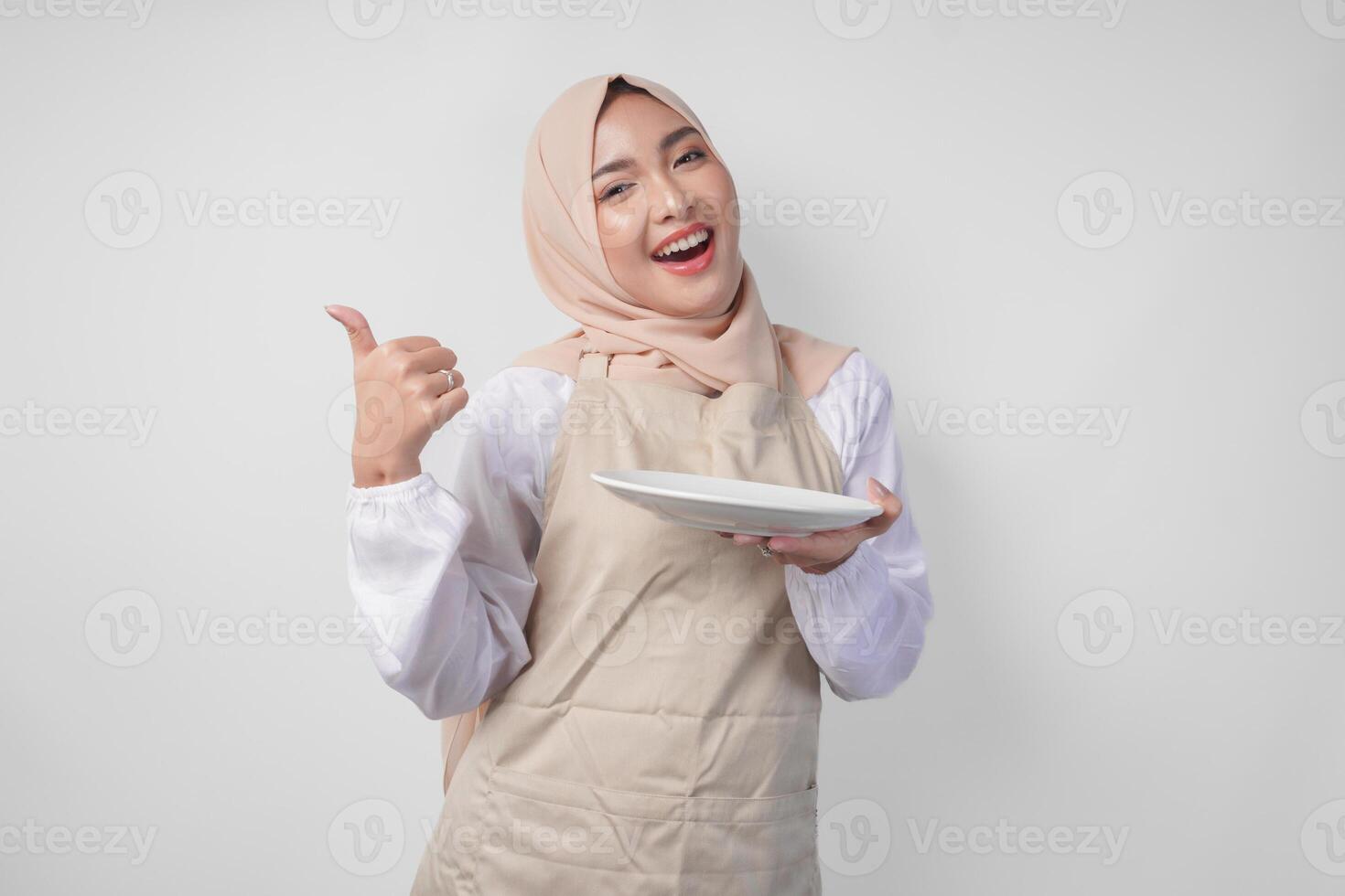 Portrait of a cheerful young Asian Muslim woman in hijab and cream apron presenting empty white plate copy space while doing thumbs up gesture photo