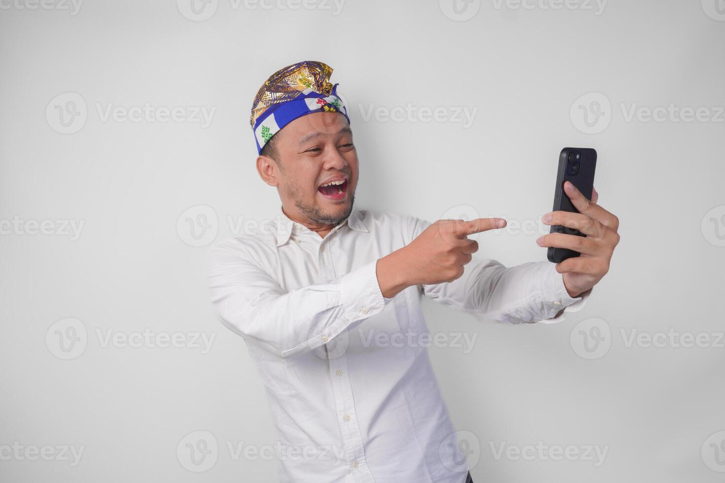 Young Balinese man in white shirt and traditional headdress doing call with family or friend feeling overjoyed and excited photo