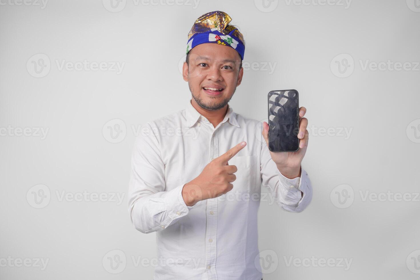 Smiling young Balinese man wearing white shirt and traditional headdress pointing to his smartphone, presenting blank screen copy space photo