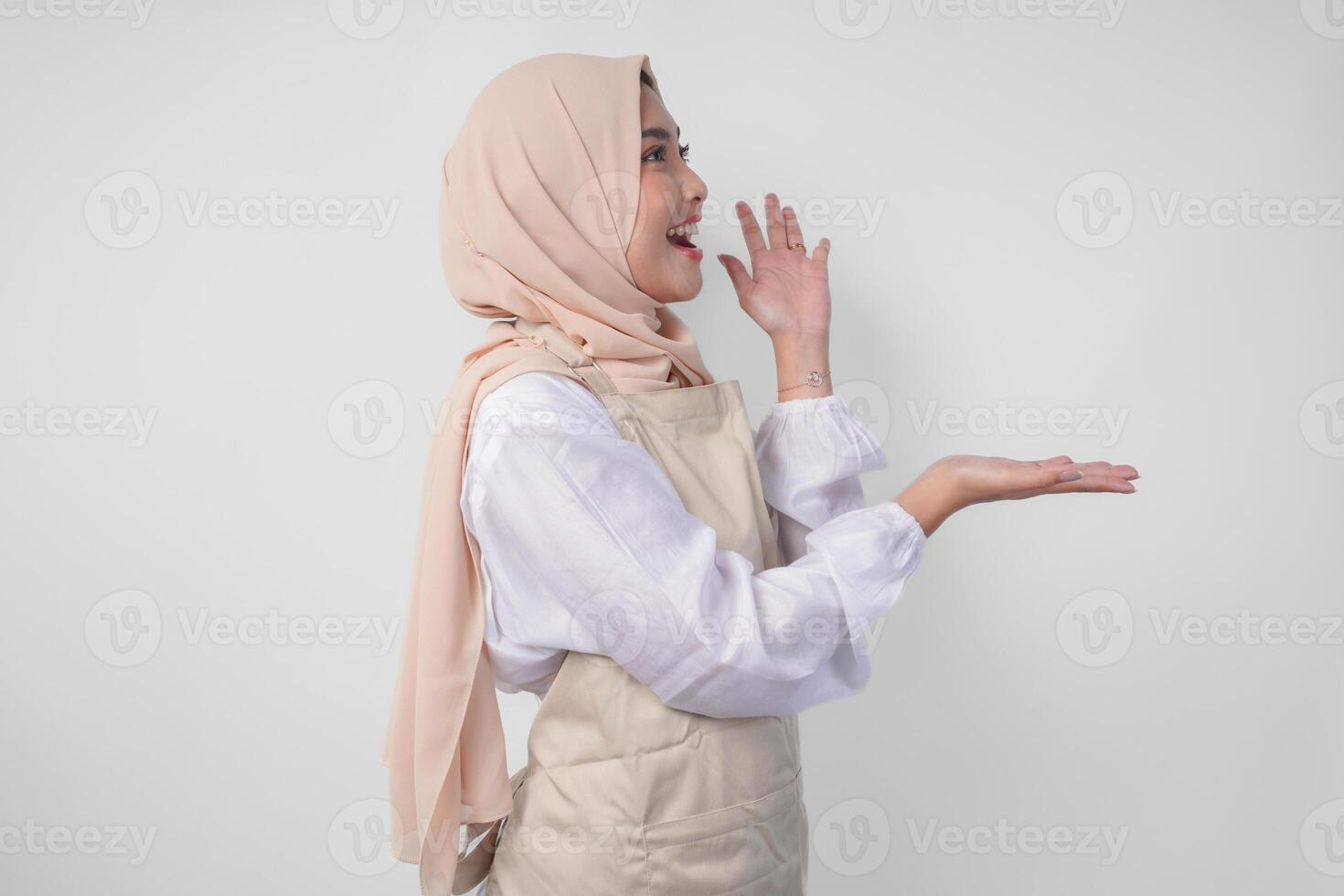 Excited young Asian Muslim woman in hijab and cream apron pointing to the empty copy space on the left and right side. Ramadan advertisement concept photo