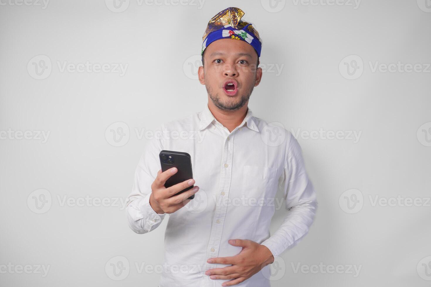 Portrait of Balinese man in white shirt and traditional headdress touching his stomach feeling hungry while using smartphone photo