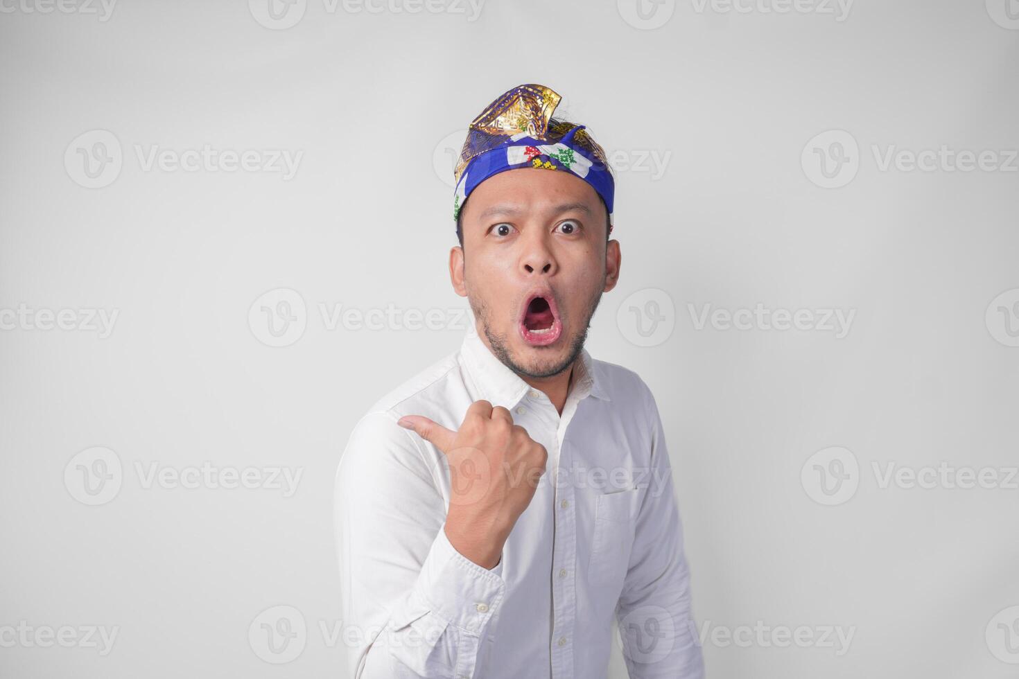 Surprised Balinese man in white shirt and traditional headdress called udeng pointing to the copy space on the left and right side photo