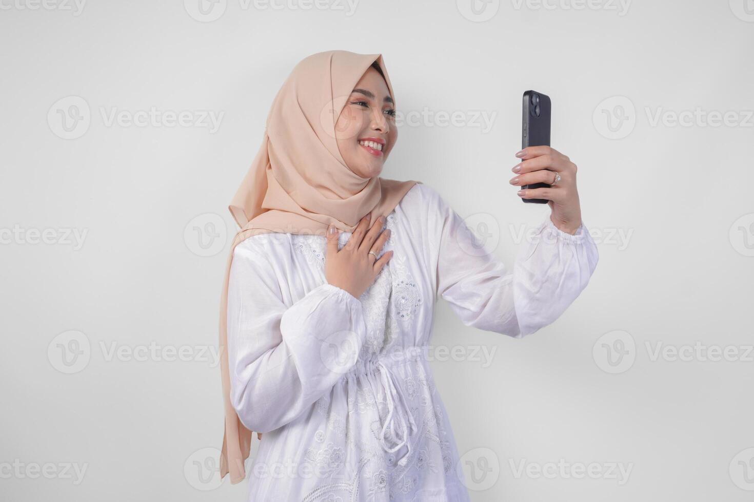 Portrait of Asian Muslim woman wearing white dress and hijab holding her smartphone to do a call to family or friend, isolated by white background. Ramadhan and Eid Mubarak concept photo