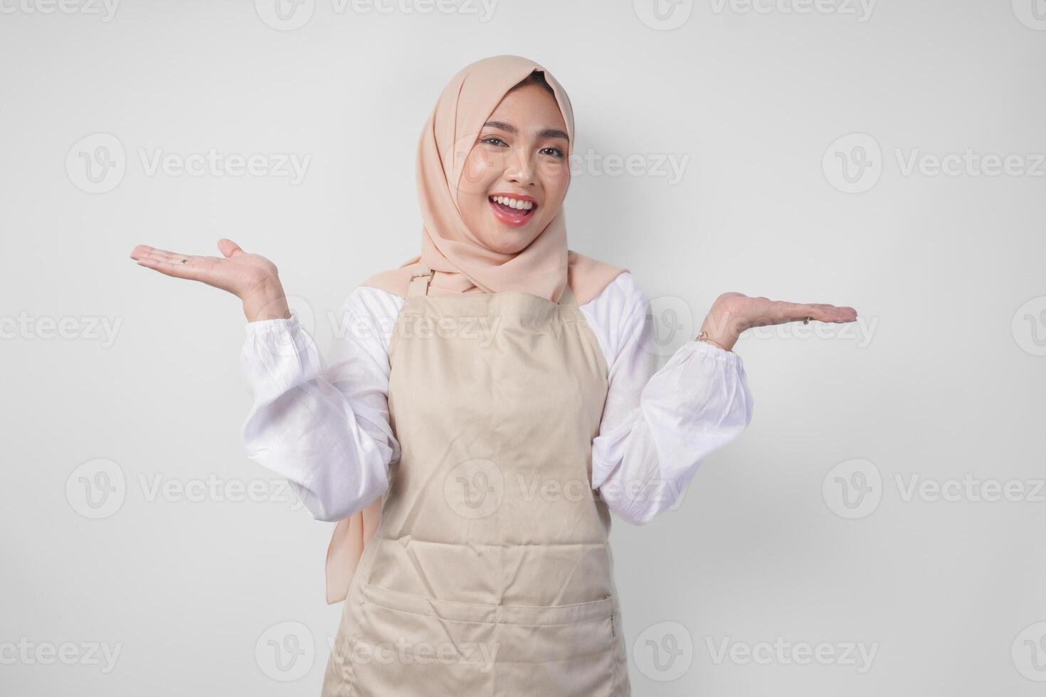 Excited young Asian Muslim woman in hijab and cream apron pointing to the empty copy space on the left and right side. Ramadan advertisement concept photo