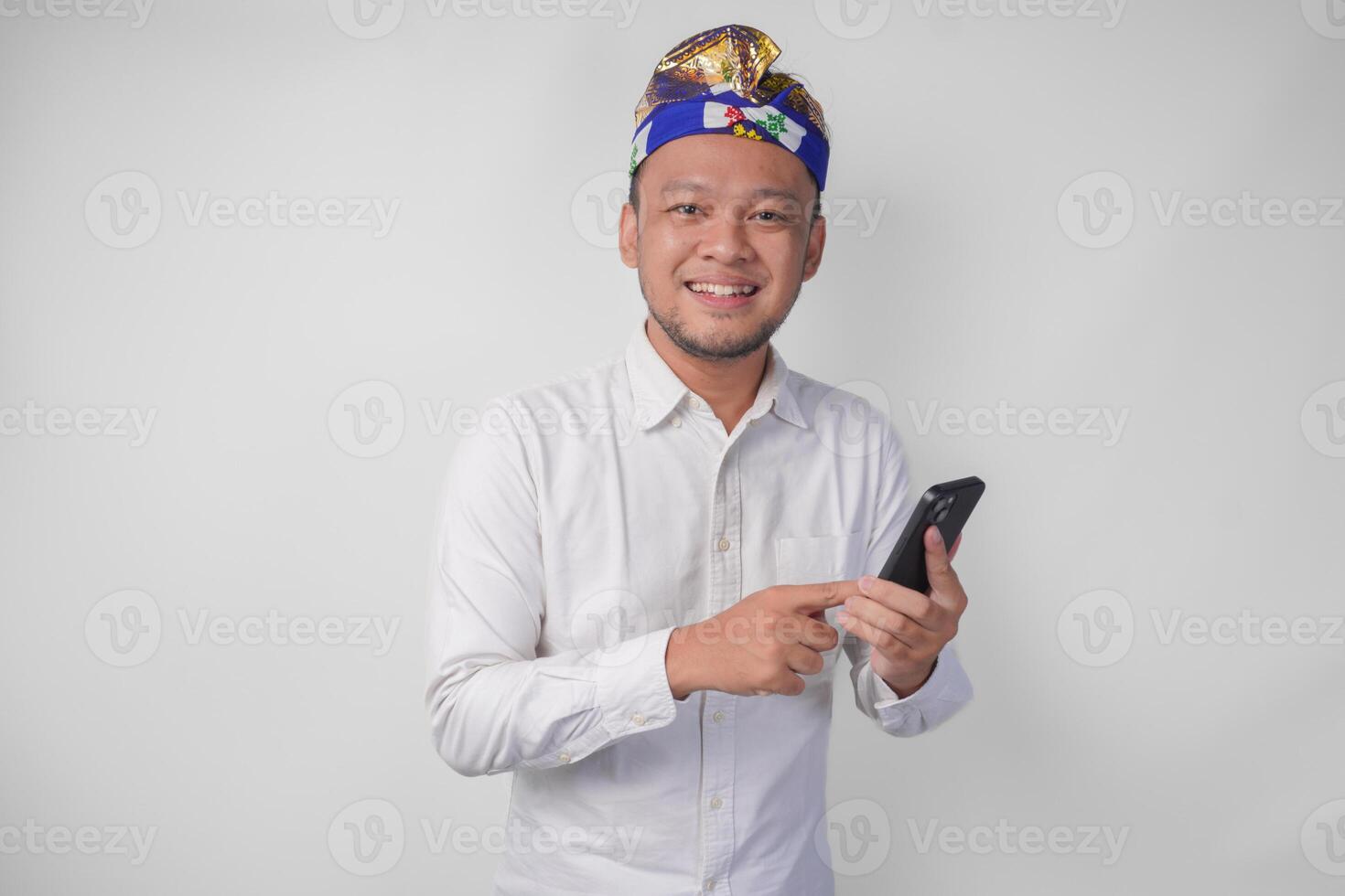 Excited Balinese man wearing white shirt and traditional headdress smiling happily while using smartphone, replying message, reading news photo
