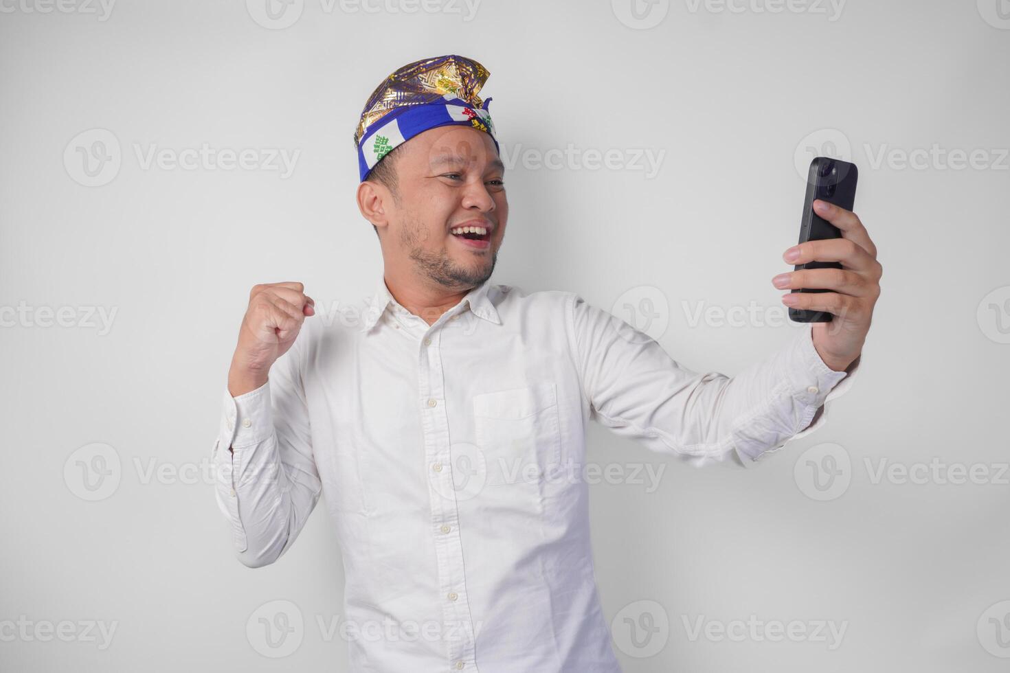 Young Balinese man in white shirt and traditional headdress doing call with family or friend feeling overjoyed and excited photo