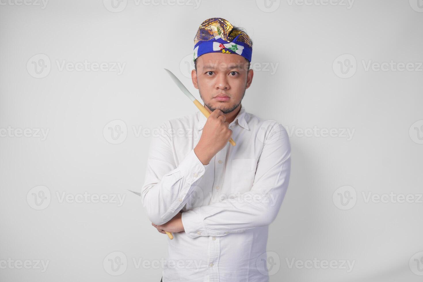Thoughtful young Balinese man in white shirt and traditional headdress holding spatula and kitchen cooking utensils while thinking hard what food menu to cook photo