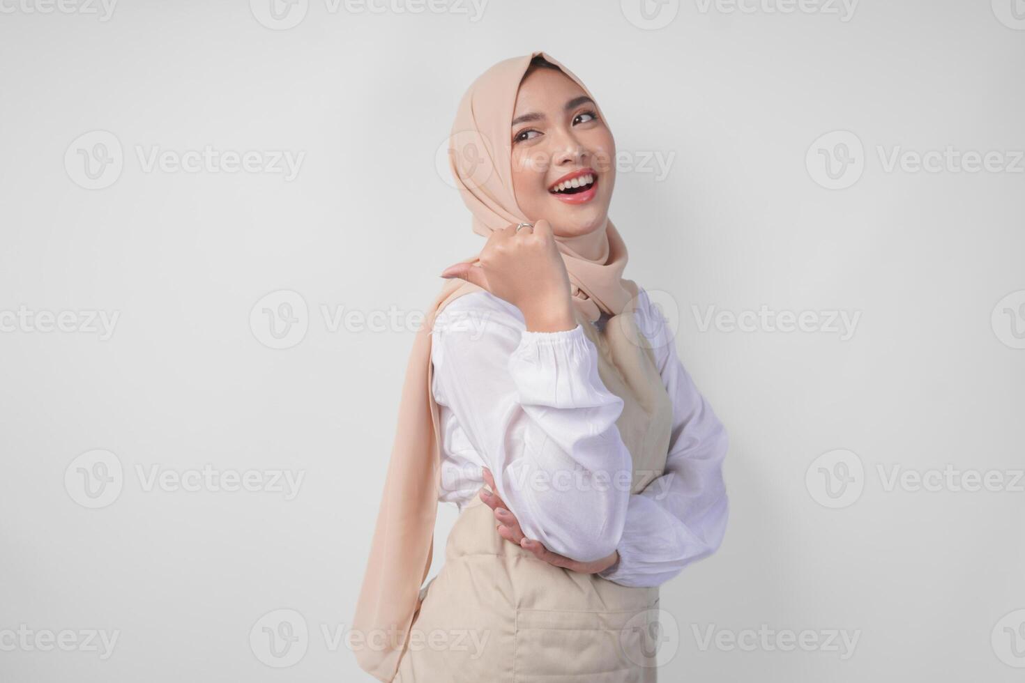 Excited young Asian Muslim woman in hijab and cream apron pointing to the empty copy space on the left and right side. Ramadan advertisement concept photo