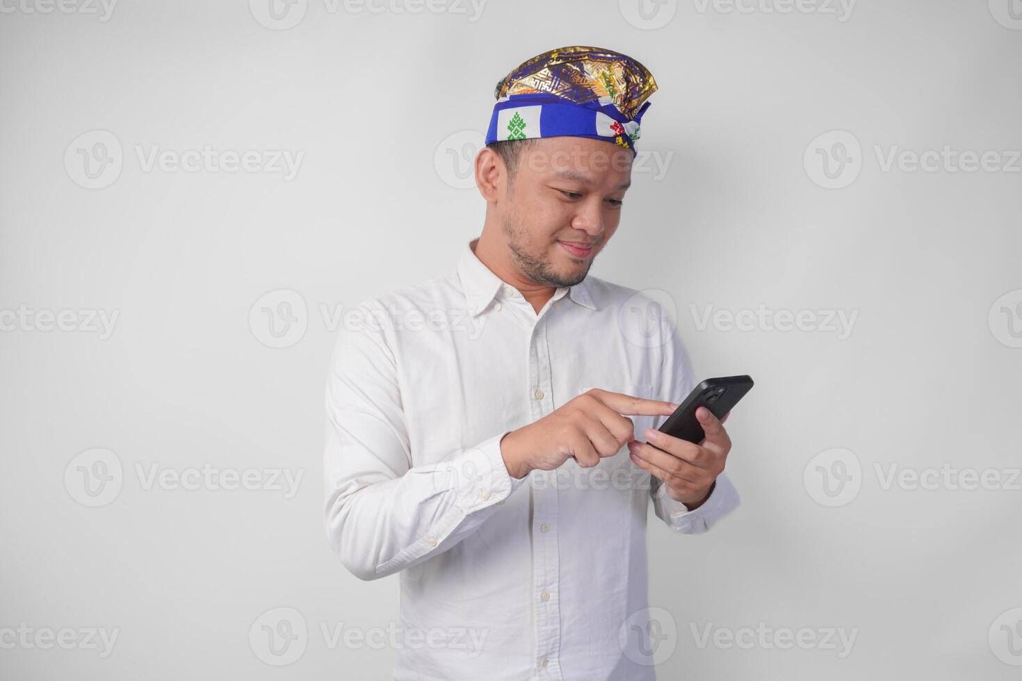 Excited Balinese man wearing white shirt and traditional headdress smiling happily while using smartphone, replying message, reading news photo