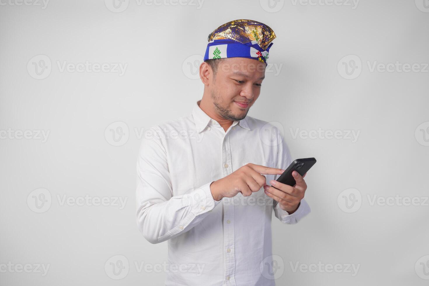 emocionado balinés hombre vistiendo blanco camisa y tradicional tocado sonriente felizmente mientras utilizando teléfono inteligente, respondiendo mensaje, leyendo Noticias foto