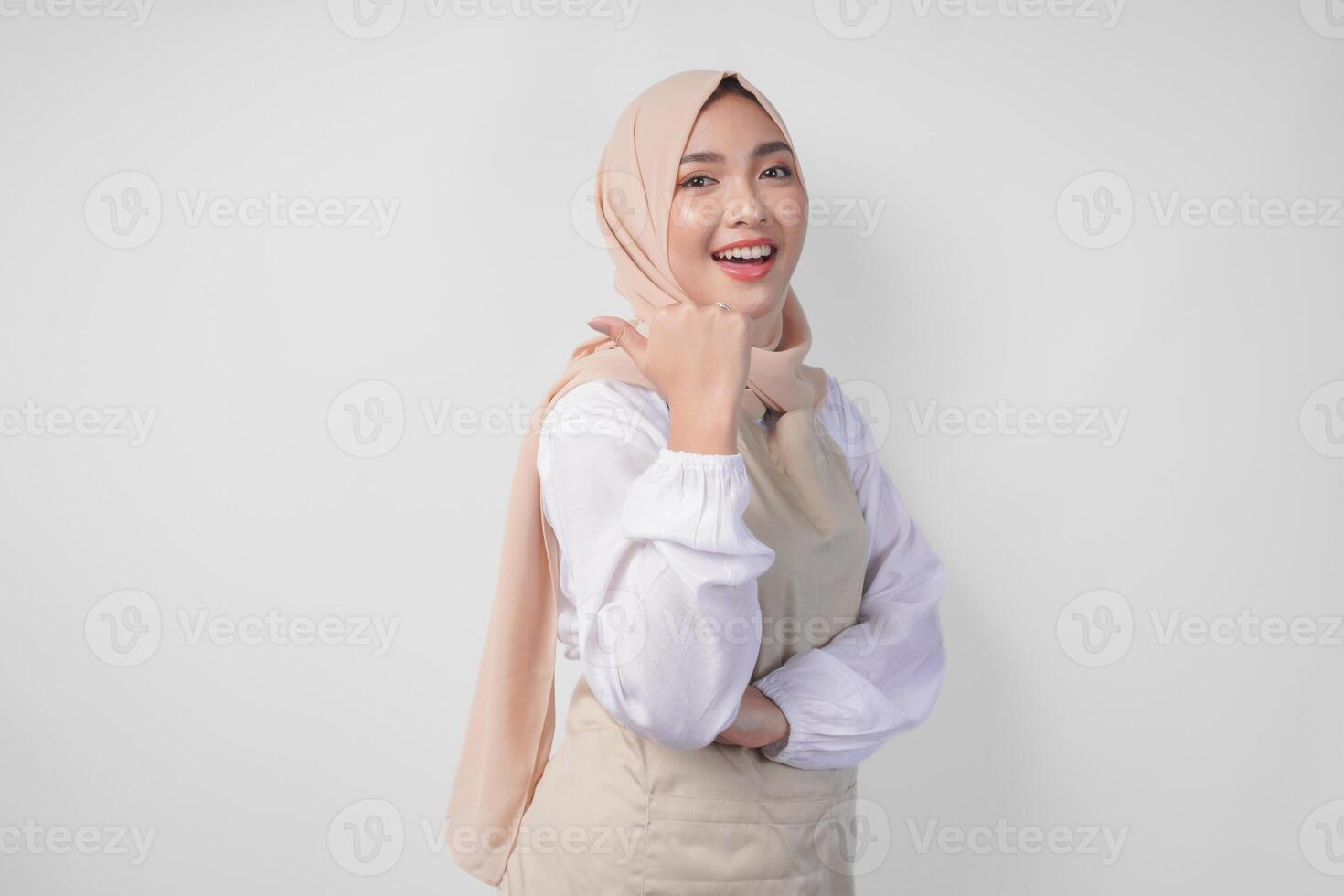 Excited young Asian Muslim woman in hijab and cream apron pointing to the empty copy space on the left and right side. Ramadan advertisement concept photo