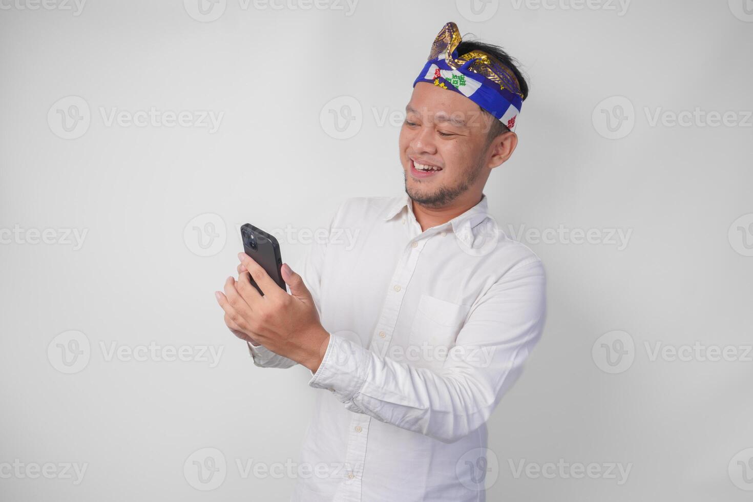 Excited Balinese man wearing white shirt and traditional headdress smiling happily while using smartphone, replying message, reading news photo