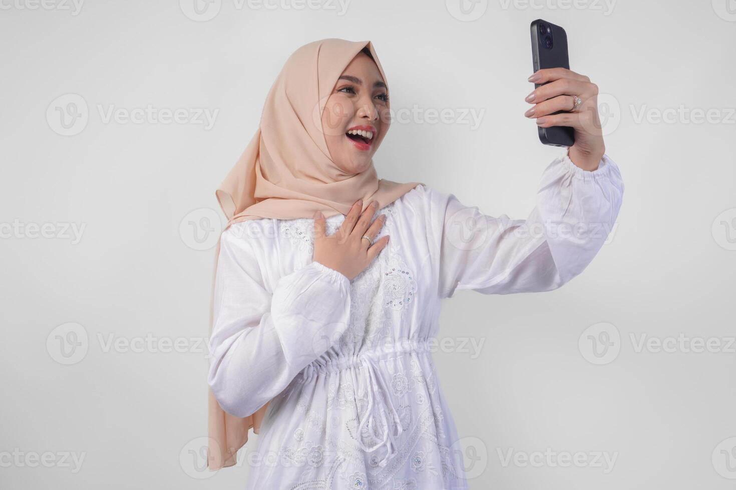 Portrait of Asian Muslim woman wearing white dress and hijab holding her smartphone to do a call to family or friend, isolated by white background. Ramadhan and Eid Mubarak concept photo