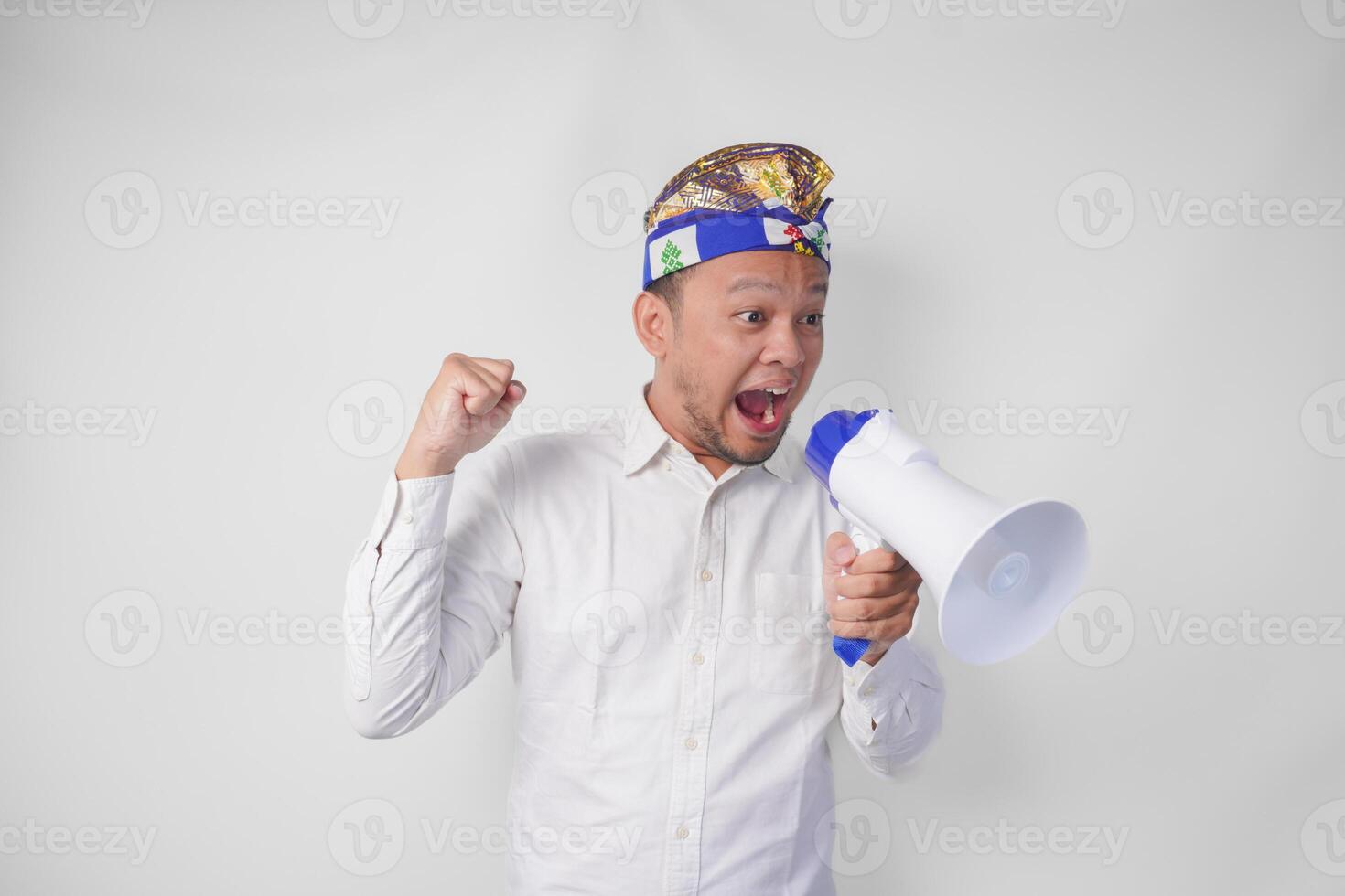 Overjoyed Balinese man in white shirt and traditional headdress shouting at megaphone feeling excited, isolated by white background photo