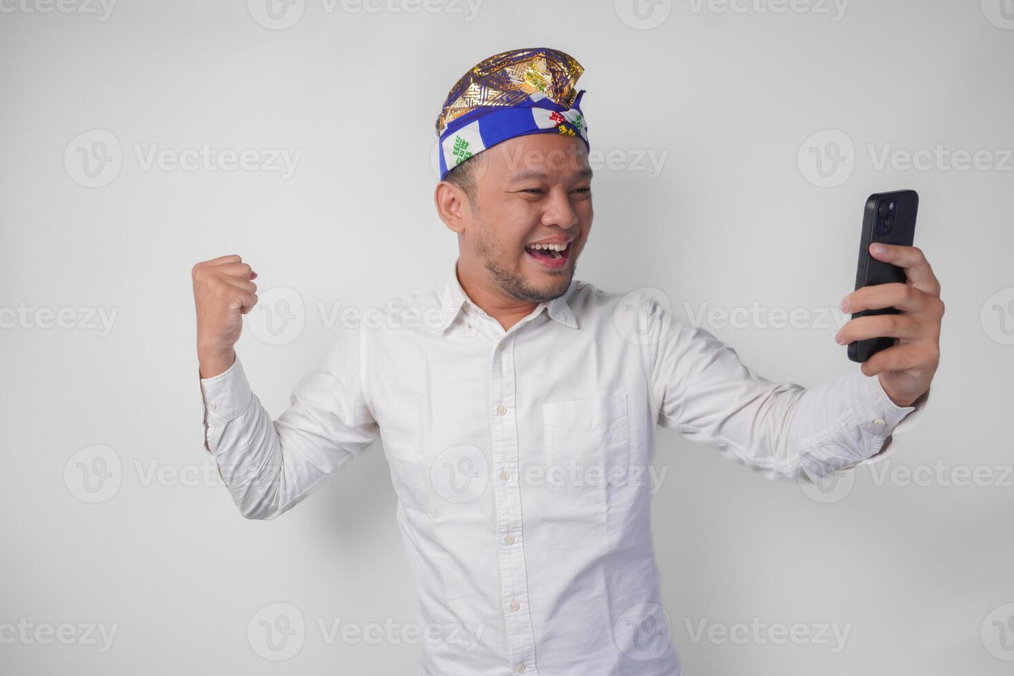 Young Balinese man in white shirt and traditional headdress doing call with family or friend feeling overjoyed and excited photo