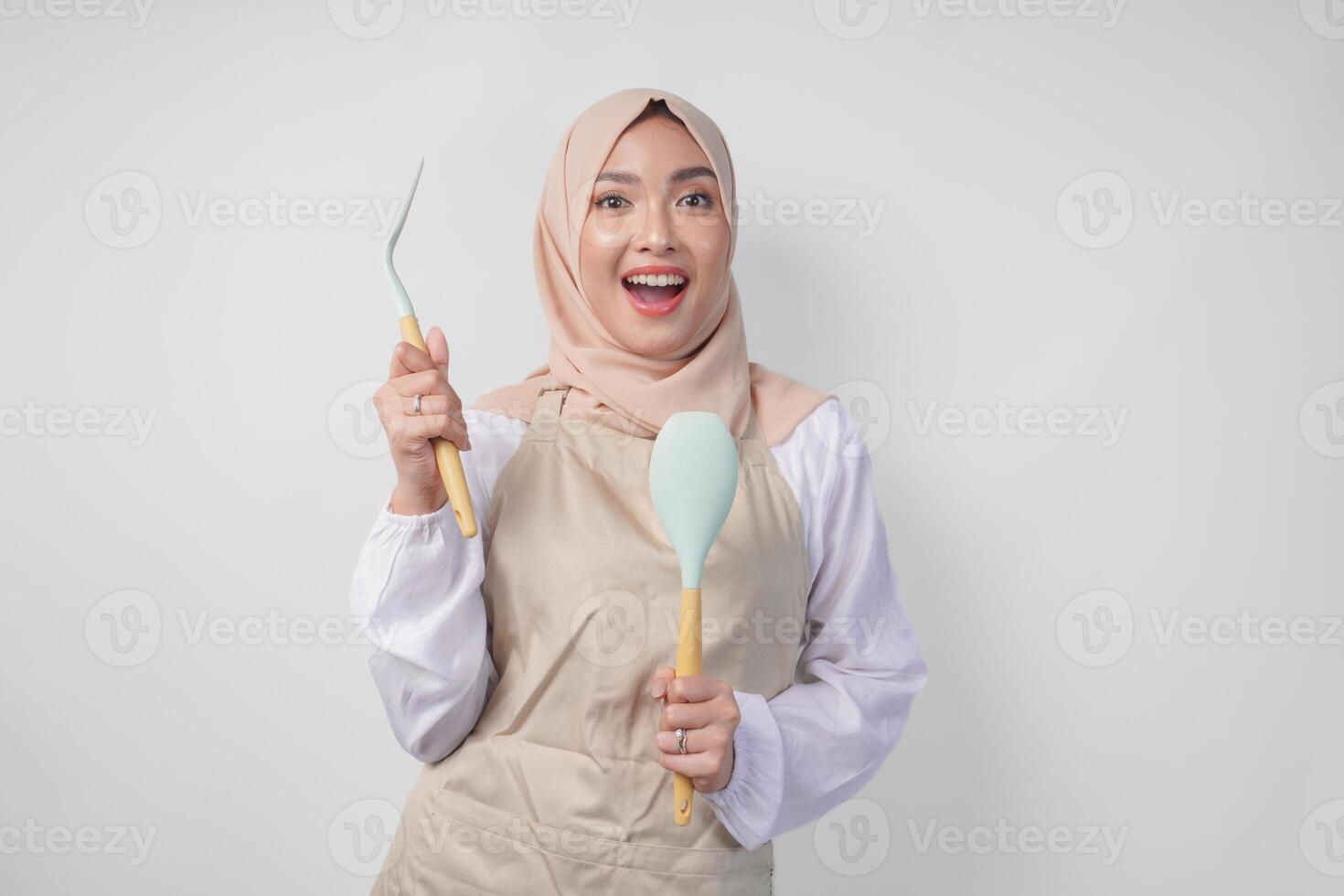 emocionado joven asiático musulmán mujer en un velo hijab y crema delantal sonriente a el cámara mientras participación espátula y cocina Cocinando utensilios foto