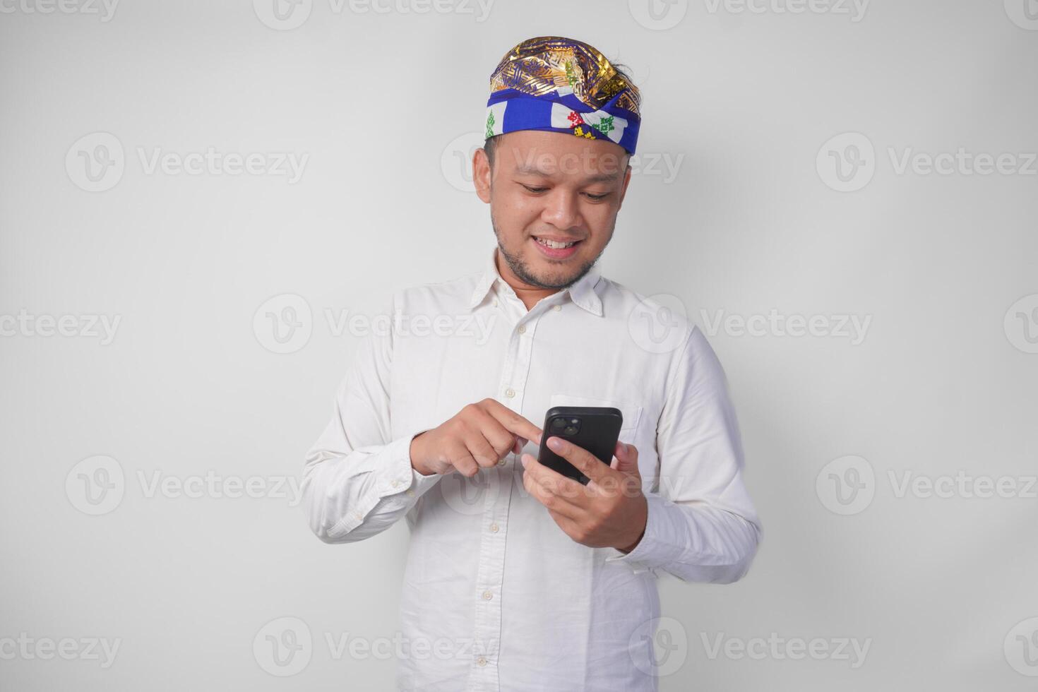 Excited Balinese man wearing white shirt and traditional headdress smiling happily while using smartphone, replying message, reading news photo