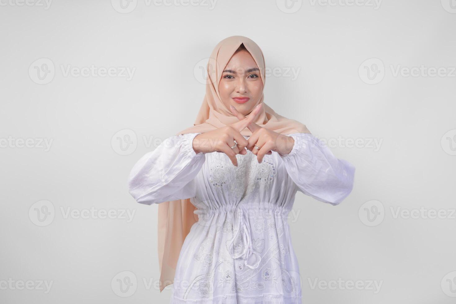 Displeased Asian woman in white dress and hijab gesturing a refusal or rejection sign, saying no, asking to stop using hands, standing over isolated white background photo