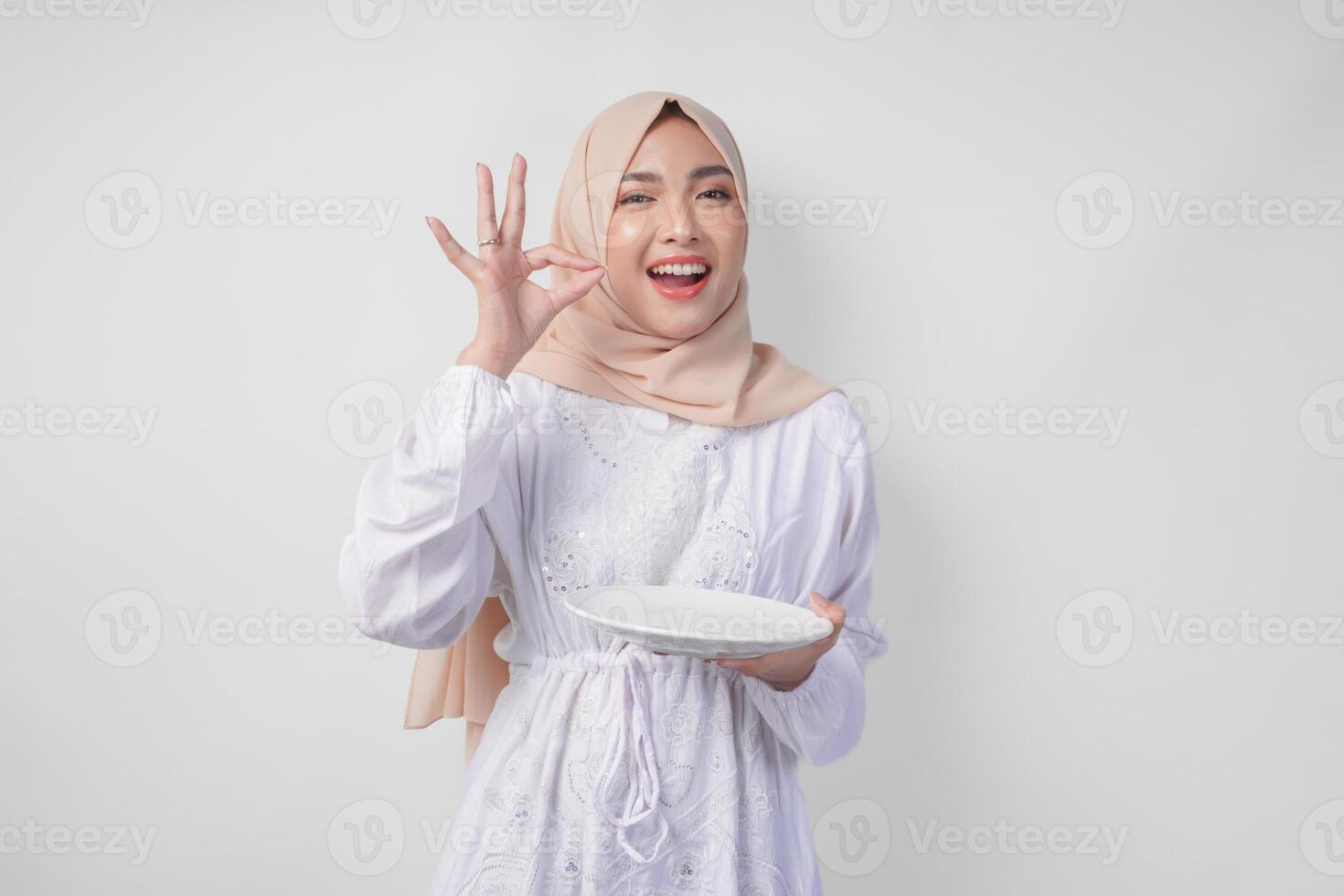 Happy young Asian Muslim woman in hijab holding an empty plate with copy space and making a delicious hand gesture to express how good the food is photo