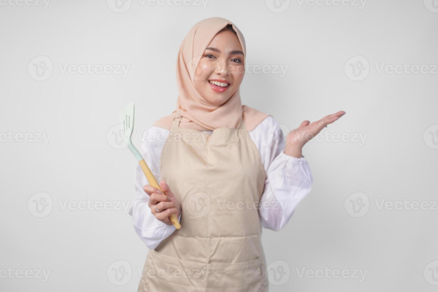 Happy young Asian Muslim woman in a veil hijab and cream apron holding spatula while pointing to the copy space beside her photo