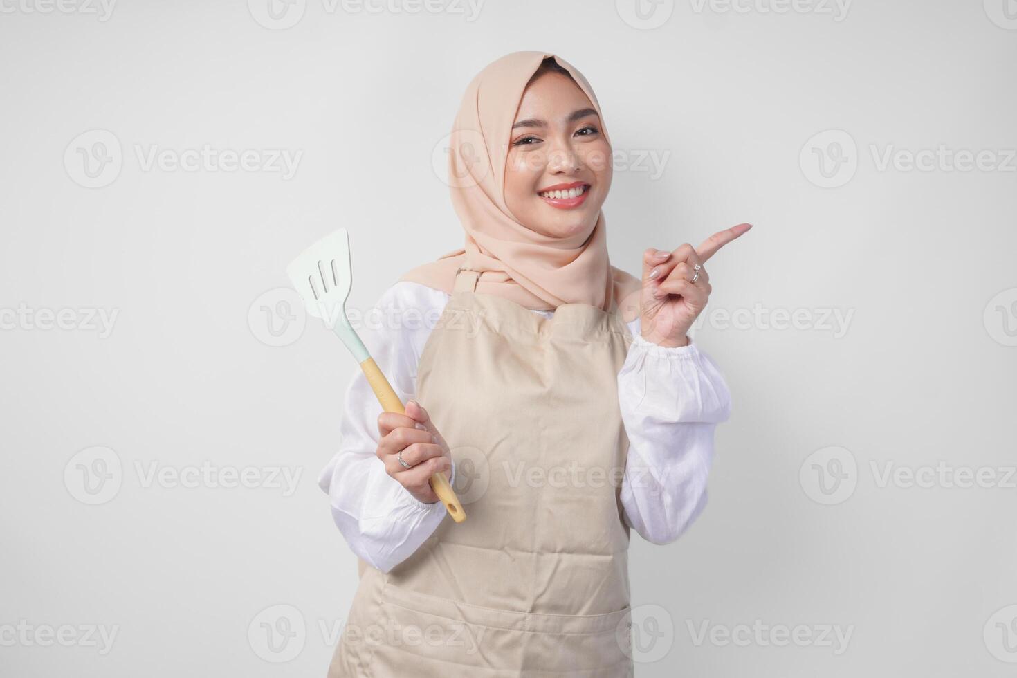 Happy young Asian Muslim woman in a veil hijab and cream apron holding spatula while pointing to the copy space beside her photo