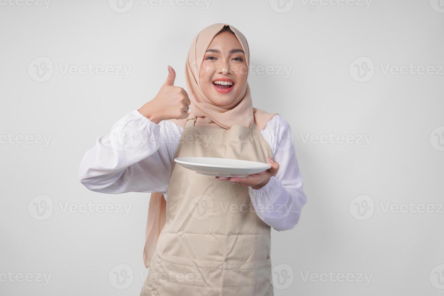 Portrait of a cheerful young Asian Muslim woman in hijab and cream apron presenting empty white plate copy space while doing thumbs up gesture photo